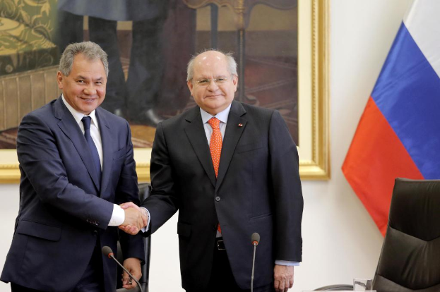 Zwei Männer in Blazern schütteln sich die Hände an Mikrofonen, mit einem Stuhl, einer Flagge und einem Wandrahmen im Hintergrund.