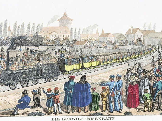 Eine Gruppe von Menschen steht um einen frühen Zug auf einem Gleis, mit einigen Passagieren im Zug und anderen auf dem Boden, vor Gebäuden und Bäumen im Hintergrund, mit der Bildunterschrift "Ludwigs-Eisenbahn, der erste Bahnhof in Deutschland" unten.