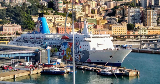 Großes Kreuzfahrtschiff an einem Stadtufer mit einer Brücke links, Fahrzeugen auf der Straße und Gebäuden und Bäumen im Hintergrund.