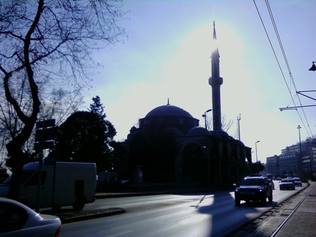 Eine Moschee steht mitten auf einer Stadtstraße, umgeben von Gebäuden, Straßenlaternen, Strommasten, Stromkabeln, Kraftfahrzeugen, Bäumen und einem klaren blauen Himmel.