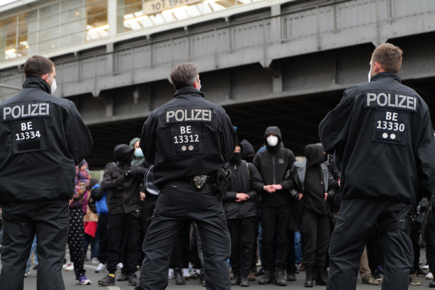 Eine Gruppe von Polizisten in Uniform steht vor einer Menge von Menschen in schwarzen Uniformen und Masken, im Hintergrund eine Brücke und ein Gebäude.
