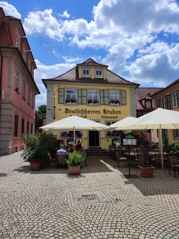Außensitzbereich mit Tischen, Sonnenschirmen und Menschen vor einem gelben Gebäude, das "Deutschen Stuben Restaurant" in Baden-Württemberg, Deutschland, unter einem blauen und weißen Himmel.