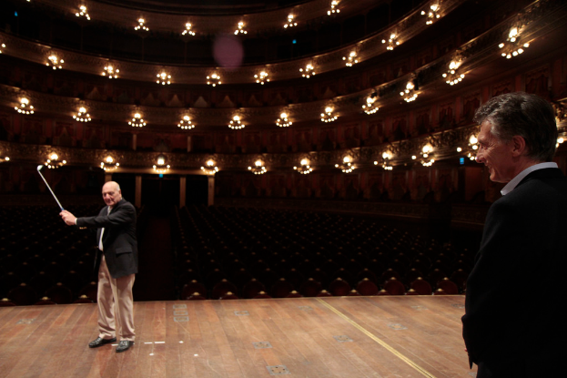 Ein älterer Mann in einem Anzug hält einen Golfschläger auf der Bühne, neben ihm steht ein weiterer älterer Mann rechts, unter Deckenlampen in einem Sitzungssaal.