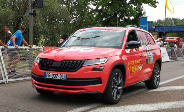 Ein rotes Auto fährt auf einer Straße neben einer Menschenmenge, einige tragen Mützen, mit Maschendrahtzaun links, einer Verkehrsampelstange, Bäumen, einer Brücke, einem Fahnenmast und einem bewölkten Himmel im Hintergrund.