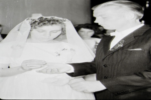 Ein Schwarz-Weiß-Foto einer Braut in einem weißen Kleid und Schleier und eines Bräutigams in einem Anzug, die sich die Eheringe reichen, mit ein paar Leuten im Hintergrund.