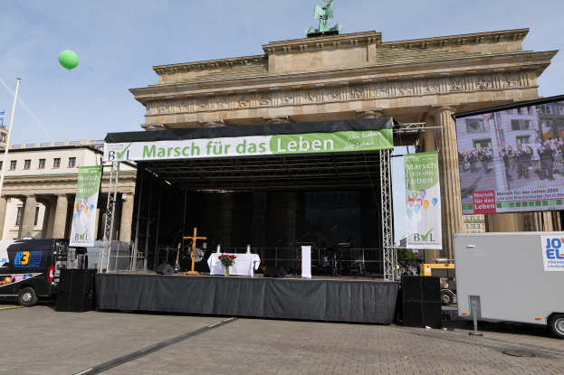 Eine Bühne vor dem Brandenburger Tor in Berlin, Deutschland, mit einem Tisch mit einer Vase, einem Kreuzsymbol, Bannern, Lautsprechern und anderen Gegenständen, sowie Fahrzeugen auf der Straße und Gebäuden im Hintergrund.