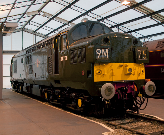 Ein Zug fährt auf den Schienen neben einem Bahnsteig mit einem am Dach befestigten Mast, der Metallstangen mit Lampen trägt.