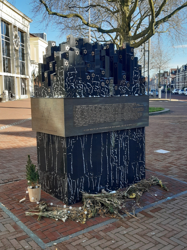 Denkmal in einem Stadtplatz für Holocaust-Opfer, mit einem Baum davor, einer Topfpflanze, verstreuten trockenen Blättern und Gebäuden, Fahrzeugen und Bäumen im Hintergrund.