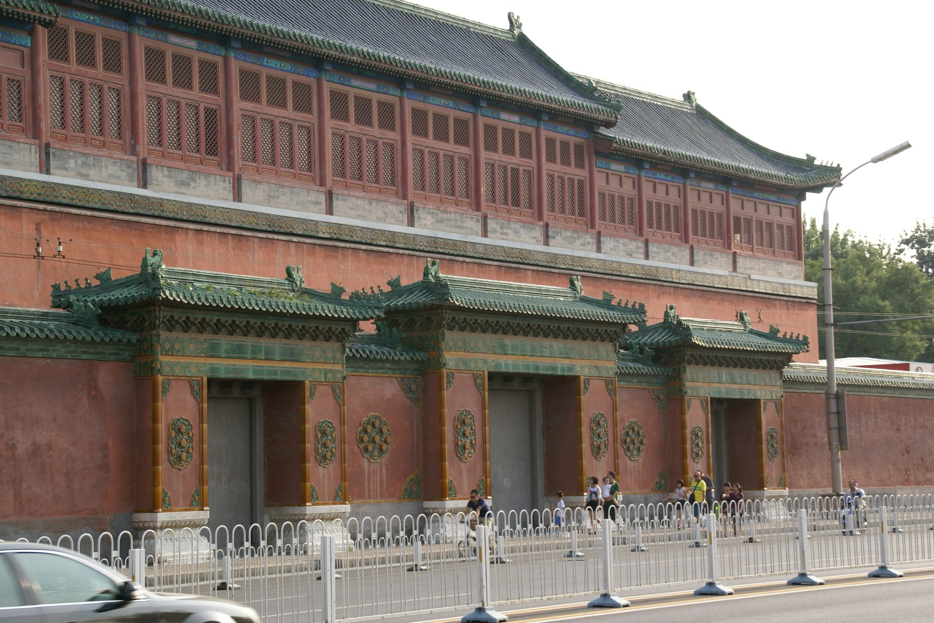 A car drives past the Forbidden City in Beijing, with pedestrians on the street, a fence, a street pole, trees, and a cloudy sky in the background.
