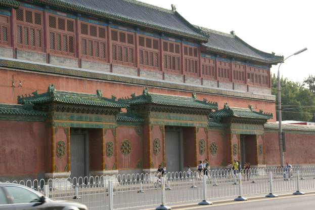 A car drives past the Forbidden City in Beijing, with pedestrians on the street, a fence, a street pole, trees, and a cloudy sky in the background.