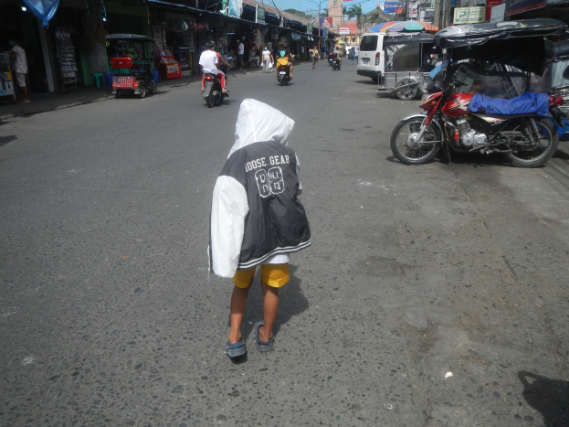 Ein junger Junge geht eine Straße voller Motorräder und anderer Fahrzeuge entlang, flankiert von Gebäuden mit Schildern, mit Bäumen, Pfählen und einem klaren blauen Himmel im Hintergrund.