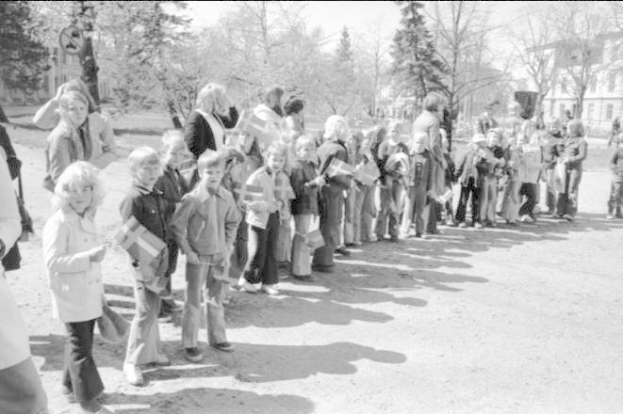Ein Schwarz-Weiß-Bild einer Gruppe von Menschen, die in einer Reihe auf einem Schotterweg stehen, Fahnen halten, mit Bäumen, Gebäuden und einem klaren Himmel im Hintergrund, die an einer Protestdemo auf dem Schulgelände teilnehmen.