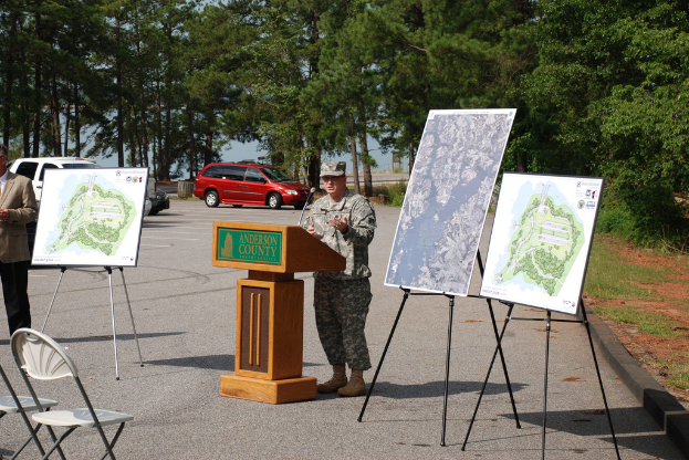 Eine Person steht an einem Podium mit einem Mikrofon, gegenüber mehreren Stühlen, mit Plakaten, Bäumen und Fahrzeugen im Hintergrund.