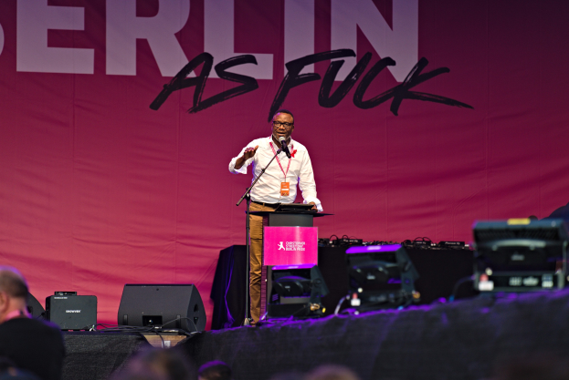 Ein Mann in einem weißen Hemd und braunen Hosen steht auf einer Bühne und hält ein Mikrofon in der Hand, während er eine Menge anspricht. Im Hintergrund ist ein pinkfarbenes Banner mit der Aufschrift 'Berlin as Fuck' zu sehen.