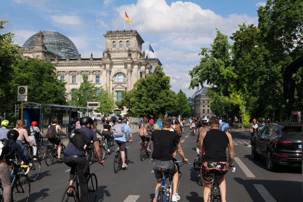 Eine Gruppe von Menschen, die auf Fahrrädern eine von Bäumen gesäumte Straße in Berlin, Deutschland, entlangfahren, mit Gebäuden auf beiden Seiten, einem Bushaltestellen auf der rechten Seite und einer Flagge, die auf einem der Gebäude weht, unter einem bewölkten Himmel.