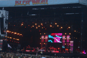 Eine Bühne mit Lichtern, einem Bildschirm und Musikern, die Instrumente spielen, mit einem Banner oben, einer Menge unten und Fahnen mit Stangen und Luftballons drumherum.