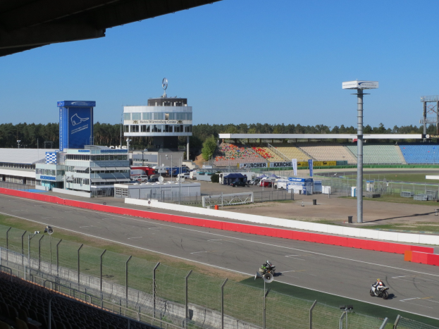 Gruppe von Motorradfahrern auf einer Rennstrecke, umgeben von einer Absperrung, Fahrzeugen, einem Stadion und Gebäuden unter einem bewölkten Himmel.