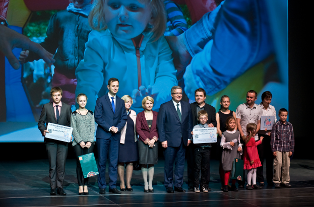 Eine Gruppe von Menschen auf einer Bühne, einige halten Taschen und Urkunden, mit einem Bildschirm im Hintergrund, der ein Kind und andere Personen anzeigt, wahrscheinlich bei einer Charity-Veranstaltung.