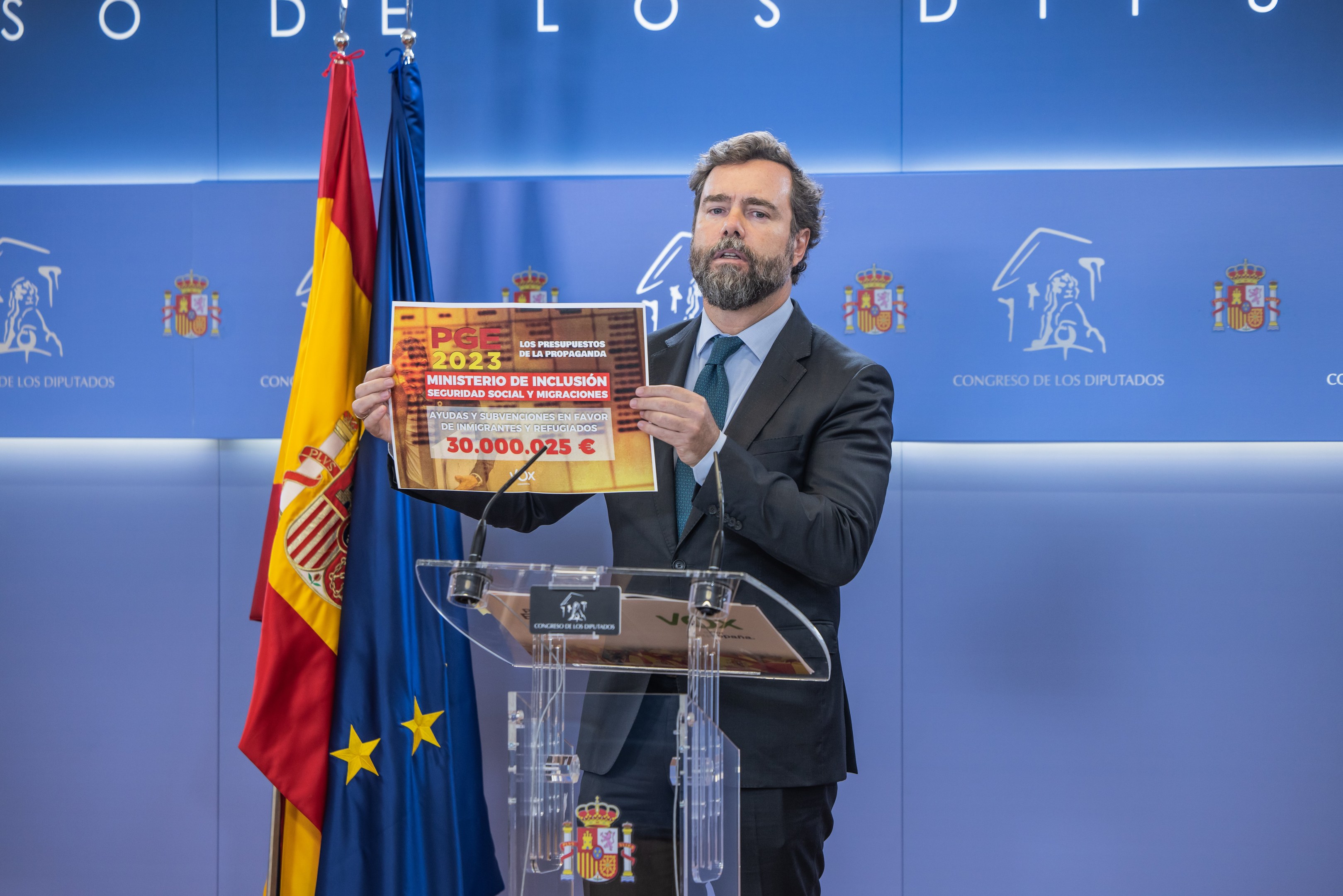 A man stands at a podium with two flags behind him, holding a book, with microphones and a board displaying text and logos.