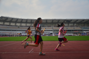 Drei Frauen auf dem Boden laufen und halten Gegenstände, mit Gruppen von Menschen, die auf einem Weg hinter ihnen stehen und einem Stadion mit Himmel im Hintergrund.