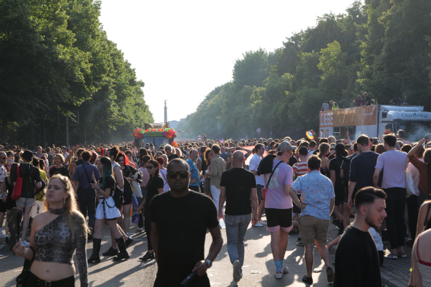 Eine große Menschenmenge, die eine von Bäumen gesäumte Straße mit einem Turm im Hintergrund und Fahrzeugen mit Teilnehmern auf der rechten Seite entlanggeht, wahrscheinlich auf dem Berliner Christopher Street Day unter einem klaren blauen Himmel.