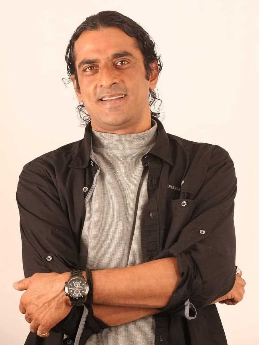 Ein Mann in einem schwarzen und aschfarbenen Anzug mit überkreuzten Armen, der eine Uhr trägt und gegen einen weißen Hintergrund steht, der tief nachdenklich erscheint.