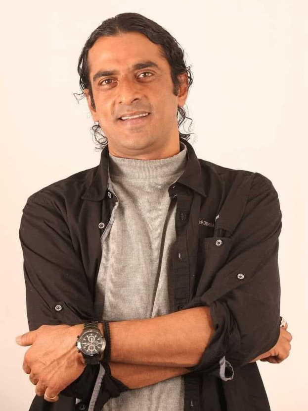 Ein Mann in einem schwarzen und aschfarbenen Anzug mit überkreuzten Armen, der eine Uhr trägt und gegen einen weißen Hintergrund steht, der tief nachdenklich erscheint.