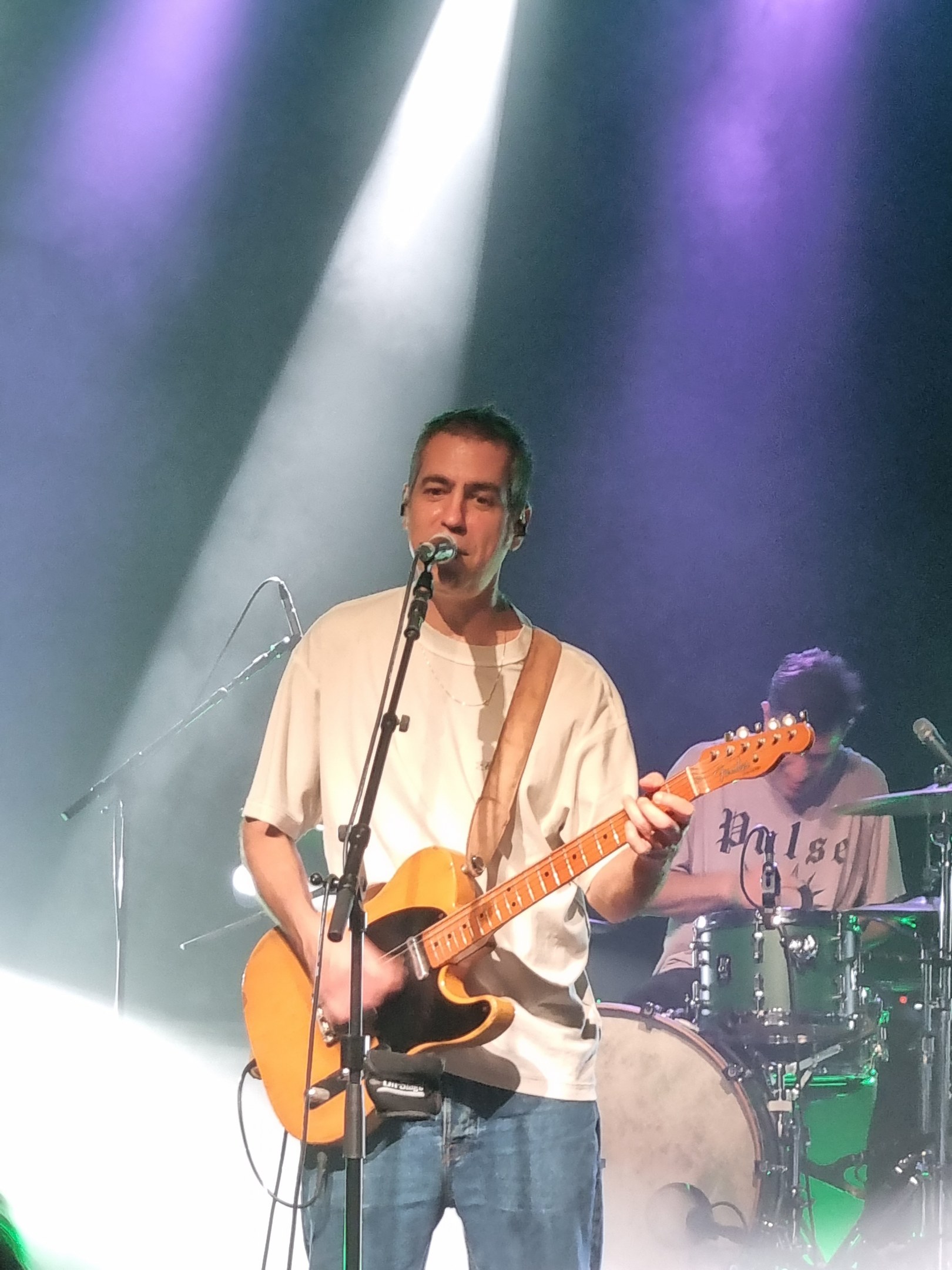 Ein Mann spielt eine E-Gitarre auf der Bühne und singt in ein Mikrofon, während ein anderer Musiker Schlagzeug spielt.