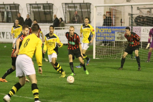 Ein Sportplatz mit mehreren Männern, die Fußball spielen, einem Netz auf der rechten Seite, einer Tafel mit Text im Hintergrund und Zuschauern, die hinter der Tafel stehen, sowie einer Wand und Fenstern im Hintergrund.