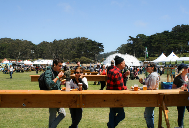 Eine Gruppe von Menschen sitzt um Tische mit Bänken, die mit Gläsern, Tellern und Essen gedeckt sind, vor einem Hintergrund aus Bäumen und Pfählen unter dem Himmel.