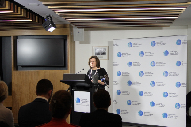 Eine Frau steht auf einer Bühne und spricht in ein Mikrofon, vor ihr sitzt ein Publikum, hinter ihr eine Wand mit einem Banner und ein Fernseher an der Holzwand, sowie Lampen an der Decke.