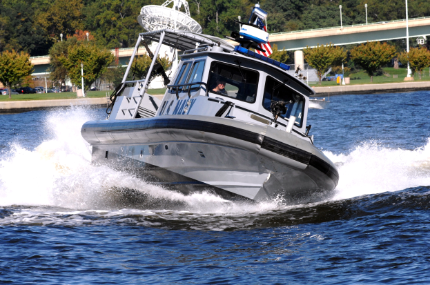 Ein Polizeiboot fährt über einen Gewässerbereich mit einer Person darin, mit einer Brücke, Bäumen, Pfählen, Fahrzeugen, Gras und einem klaren blauen Himmel im Hintergrund.