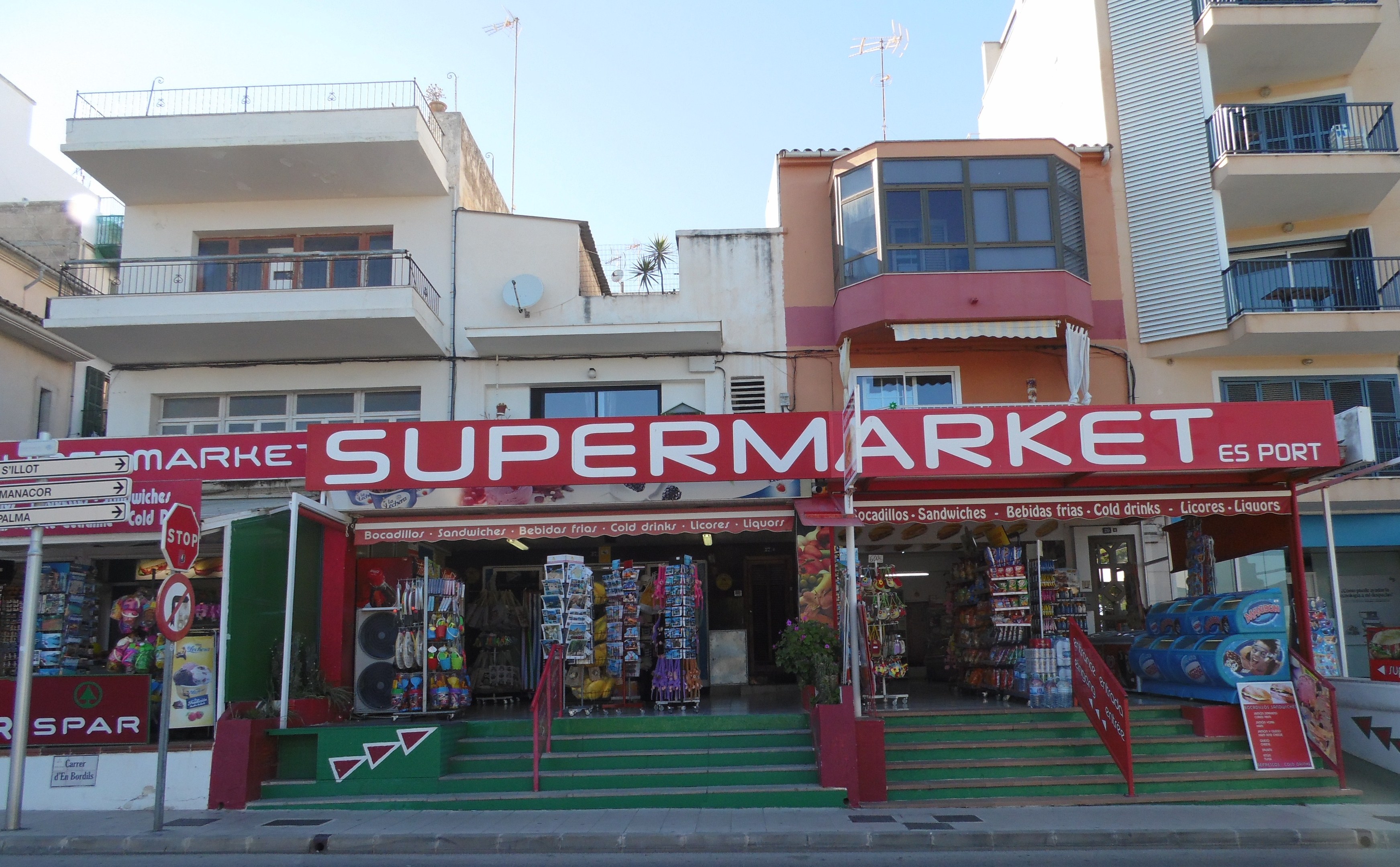 A supermarket at a street corner with buildings in the background, featuring various foreground elements like store signs, poles, plants, and a visible sky above.