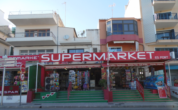 A supermarket at a street corner with buildings in the background, featuring various foreground elements like store signs, poles, plants, and a visible sky above.