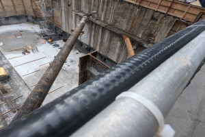 Eine Baustelle mit einem großen Metallrohr in der Mitte, umgeben von Arbeitern, hölzernen Gegenständen und verschiedenen Geräten, mit Fahrzeugen und einem Gebäude im Hintergrund.