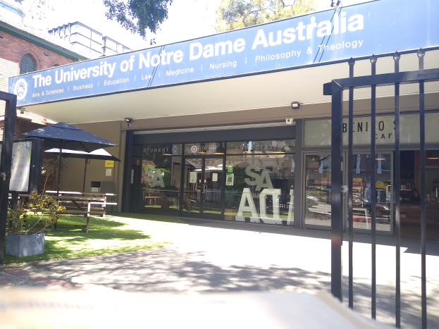 Das Gebäude der University of Notre Dame Australia mit Glaswänden und -türen, einem Schild, einem Metallzaun, Gras, Topfpflanzen, Bänken, einem Außenständer, einer Baumgruppe und einem bewölkten Himmel.