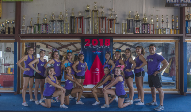 Gruppe von Cheerleadern in lila T-Shirts und schwarzen Shorts, die vor einer Vitrine mit Trophäen und einem Banner '2018 Summit Cheerleading Champions' posieren.