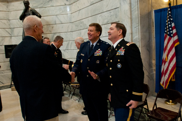 Menschen schütteln sich die Hände vor Stühlen, mit einer Statue, einer Fahne und einem Vorhang im Hintergrund.