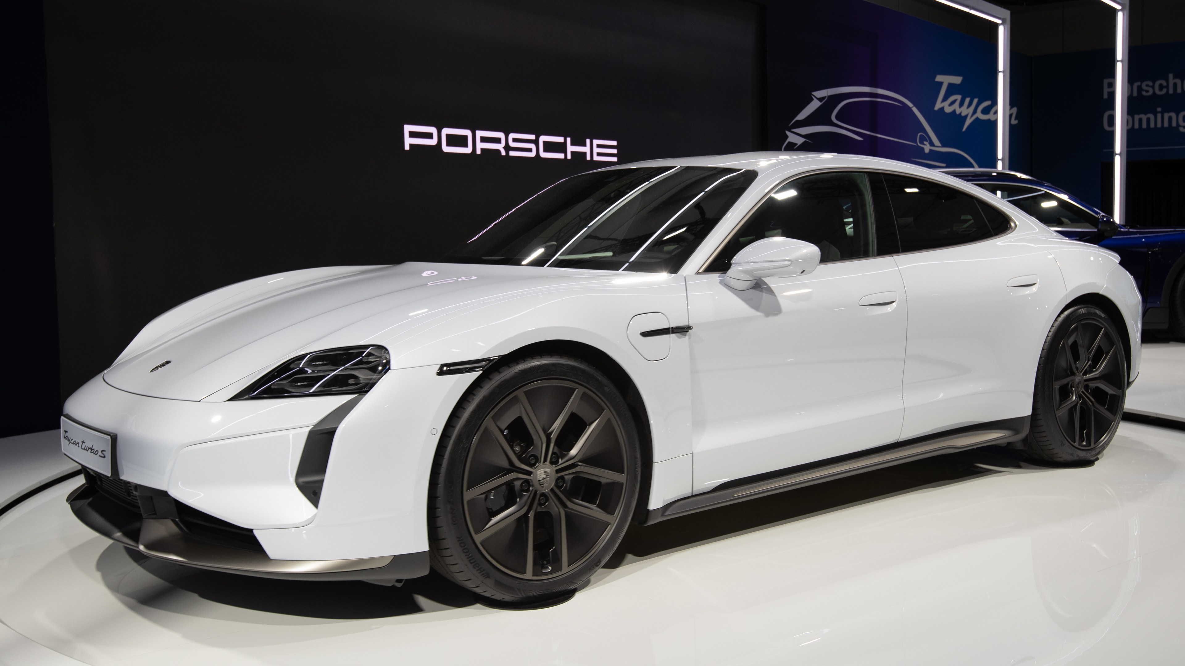 Weißer Porsche Taycan Turbo S E-Hybrid auf der IAA in Frankfurt, umgeben von schwarzen Schlafzimmermöbeln in einem modernen Wohnsetting.
