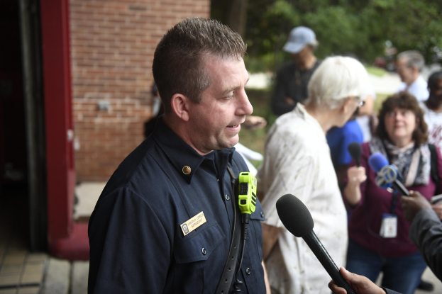 Polizeibeamter spricht mit Reportern mit Mikrofonen außerhalb bei Bäumen und einem Gebäude.