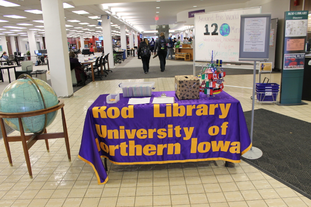 Eine Ausstellung mit einem Tisch, auf dem verschiedene Landesflaggen, eine Box, Papiere und ein Banner stehen, eine Kugel links daneben, ein Anschlagbrett rechts daneben und mehrere Tische und Stühle in der Ferne.