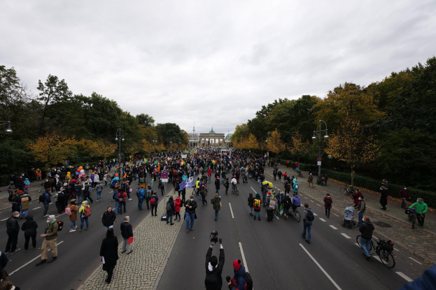 Eine große Gruppe von Menschen geht eine von Bäumen gesäumte Straße in Berlin entlang, einige halten Kameras, während sie an einer Protestdemo mit einem Gebäude und einem klaren Himmel im Hintergrund teilnehmen.