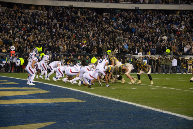 Fußballspieler in Sportkleidung und Helmen liegen auf dem Boden während eines Spiels in einem Stadion mit Zuschauern, Geländern und Tafeln im Hintergrund.