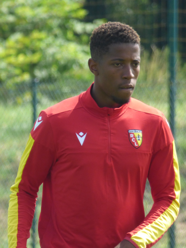 Ein junger Mann in einem roten und gelben Trikot steht auf einem Fußballfeld, umgeben von einem Zaun und Bäumen, mit einem entschlossenen Gesichtsausdruck, wahrscheinlich als Flügelspieler.