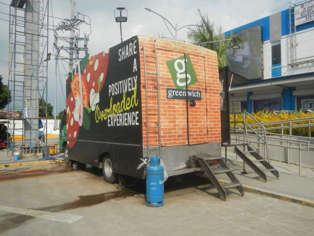 Ein weißer Lastwagen vor einem Gebäude mit einer Tafel, auf der "Teilen Sie eine positiv überladene Erfahrung" steht, mit Stufen, einem Zylinder auf dem Boden, Geländern, Strommasten mit Drähten, Laternenmasten, Bäumen und einem bewölkten Himmel im Hintergrund.