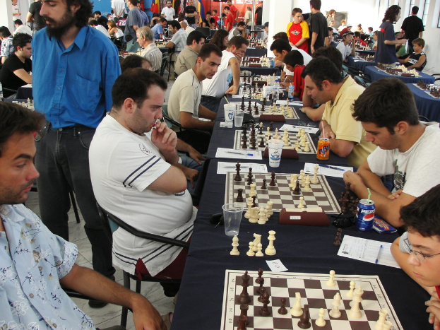 Eine Gruppe von Menschen spielt Schach mit einem Mann in einem blauen Hemd auf der linken Seite, ein Fenster im oberen rechten Eck und ein Tisch mit einem Glas, Schachbrett, Schachfiguren, einer Coke, Papier, Stift und einer Tischdecke.