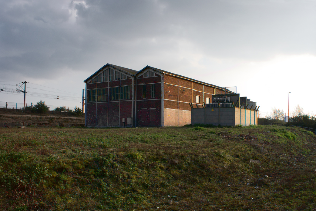 Eine Szene mit Gartenhütten, Transformatoren, Strommasten, Kabeln, Zäunen, einer Straßenlaterne, Bäumen und einem bewölkten Himmel.