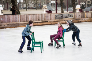 Kinder beim Skifahren vor einem Spielplatz, mit drei Kindern und zwei Stühlen in der Mitte und Gebäuden, Bäumen, Bänken, Pfosten und einem Basketballfeld im Hintergrund.