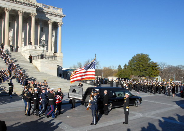 Funeral procession with people in caps and flags outside the U.S. Capitol Building, including vehicles, steps, railings, light poles, trees, and sky.