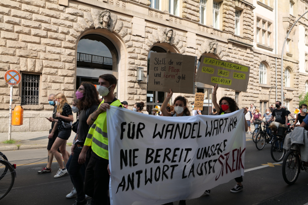 A group of people walking down a street during a protest in Berlin, some holding placards and banners, others riding bicycles, with a building featuring windows, arches, pillars, and sculptures in the background.
